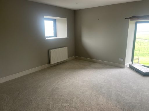 View of a large room with a grey carpet. There is a small window and a large windowed door looking out to the garden